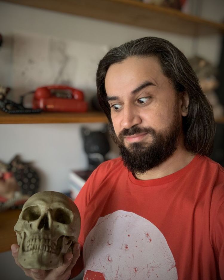 otografia de meio corpo do Dr. Bruno Marinho de Sousa, psicólogo e pesquisador, sorrindo de forma irônica enquanto segura uma caveira humana decorativa em um escritório com estante de livros ao fundo.